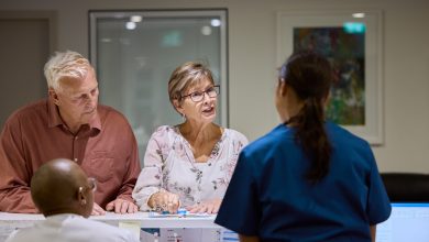 Elderly couple talking to medical staff at hospital reception desk. Conversation seems focused and attentive. Bright, organized setting reflects professional healthcare environment.
