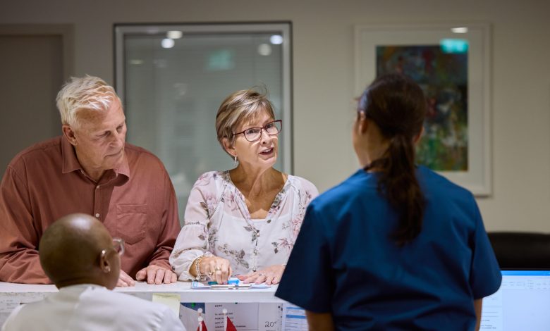 Elderly couple talking to medical staff at hospital reception desk. Conversation seems focused and attentive. Bright, organized setting reflects professional healthcare environment.