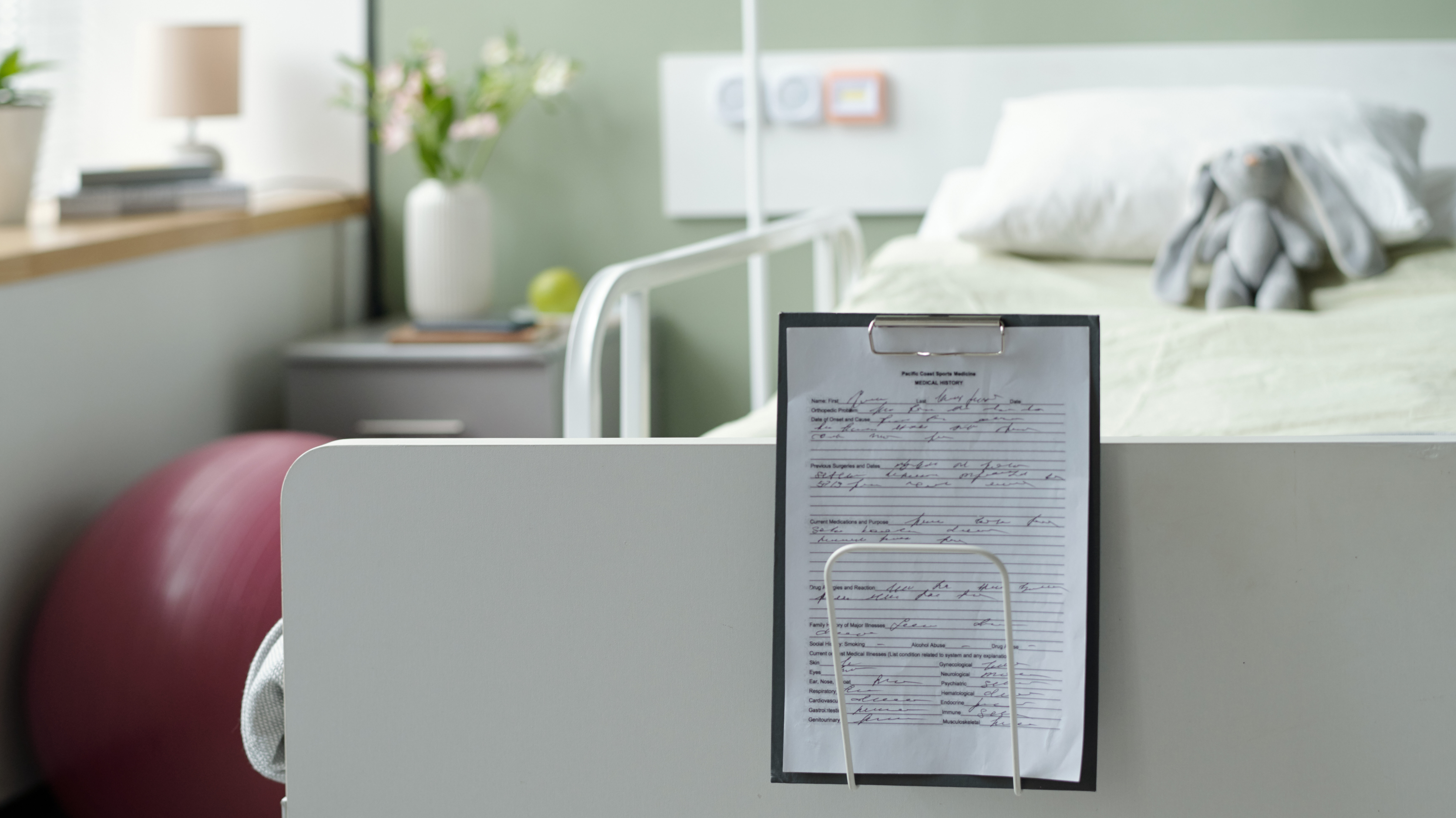 Medical bed in hospital room with clipboard holding handwritten notes in foreground and exercise ball in background with vases and medical equipment