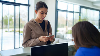 A woman holding her smartphone while engaging in conversation with a healthcare worker at the clinic’s front desk. The scene suggests appointment confirmation, digital check-in, or accessing medical information through mobile technology in a modern healthcare setting.