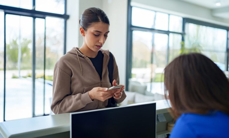 A woman holding her smartphone while engaging in conversation with a healthcare worker at the clinic’s front desk. The scene suggests appointment confirmation, digital check-in, or accessing medical information through mobile technology in a modern healthcare setting.