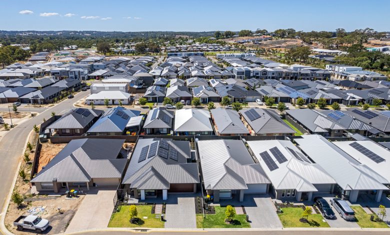 Low aerial close view new dense rural housing development, mostly grey roofing, some green landscaping, young trees. Rows of houses of various architectural styles with shades of grey roofing in rural urban sprawl, some still under construction. Tightly-packed houses with small front and back yards and few if any trees. Some solar panels installed. Mt Barker near Adelaide, South Australia