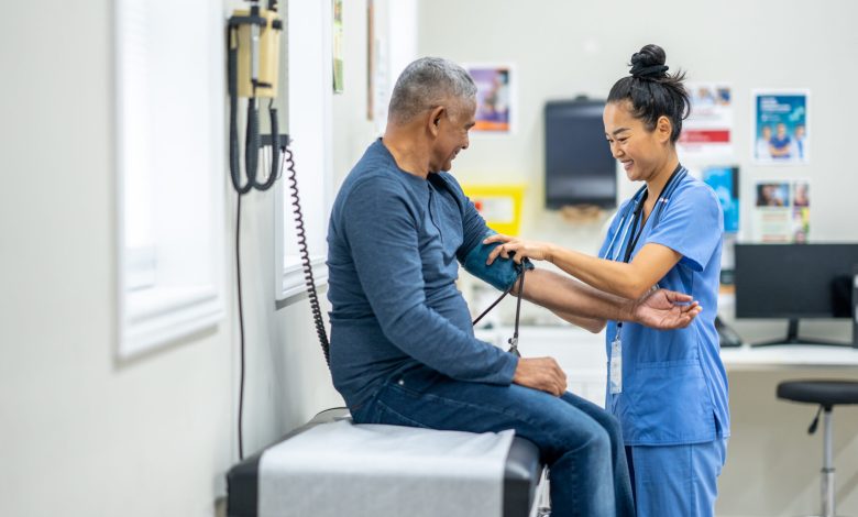 A senior gentleman of Hispanic decent, sits up on an exam table as his female nurse takes his blood pressure. The nurse is wearing blue scrubs and making small talk to put the gentleman at ease.