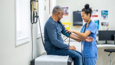 A senior gentleman of Hispanic decent, sits up on an exam table as his female nurse takes his blood pressure. The nurse is wearing blue scrubs and making small talk to put the gentleman at ease.