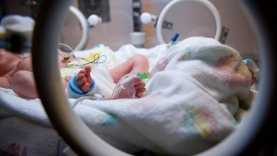 Looking through the access port on an Isolette at the foot of a premature infant with IV line and medical tape.
