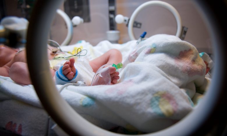 Looking through the access port on an Isolette at the foot of a premature infant with IV line and medical tape.