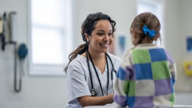 A female Paediatrician sits in front of a young patient on her exam table as she gives her a check-up. The doctor is wearing white scrubs and is smiling to put the young girl at ease.