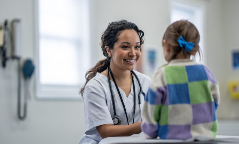 A female Paediatrician sits in front of a young patient on her exam table as she gives her a check-up. The doctor is wearing white scrubs and is smiling to put the young girl at ease.