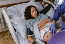 A pregnant multiracial woman lies in a hospital bed and lifts up her hospital gown as a nurse makes adjustments to the monitors on her abdomen which are tracking contractions and the baby's heartbeat.