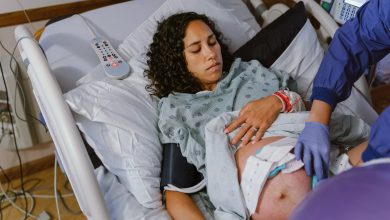 A pregnant multiracial woman lies in a hospital bed and lifts up her hospital gown as a nurse makes adjustments to the monitors on her abdomen which are tracking contractions and the baby's heartbeat.
