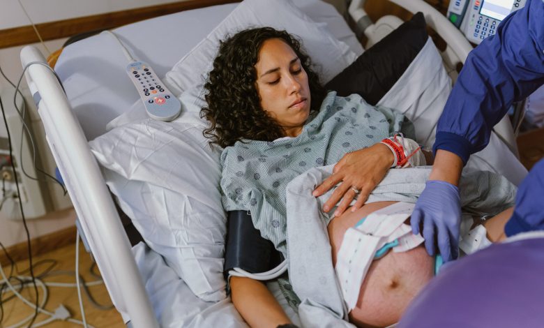A pregnant multiracial woman lies in a hospital bed and lifts up her hospital gown as a nurse makes adjustments to the monitors on her abdomen which are tracking contractions and the baby's heartbeat.