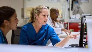 Nurse pointing at computer screen while discussing patient care with colleague. Medical staff working together in busy hospital setting. Healthcare professionals focused on providing best treatment.