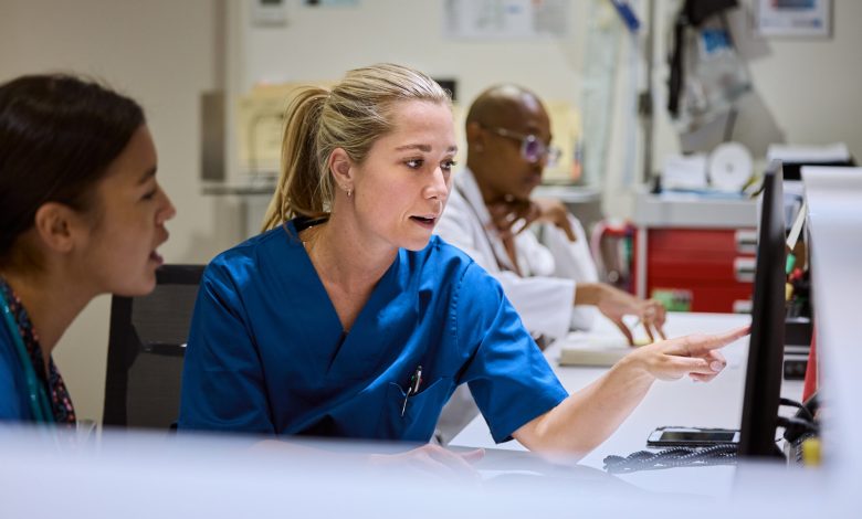 Nurse pointing at computer screen while discussing patient care with colleague. Medical staff working together in busy hospital setting. Healthcare professionals focused on providing best treatment.