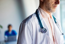 Male doctor wearing a stethoscope and white coat stands in a clinic with a blurred colleague behind him.
