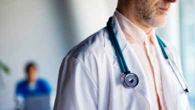 Male doctor wearing a stethoscope and white coat stands in a clinic with a blurred colleague behind him.
