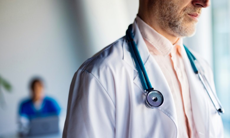 Male doctor wearing a stethoscope and white coat stands in a clinic with a blurred colleague behind him.