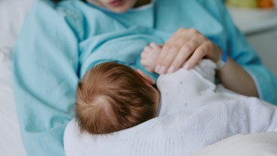 Mother breastfeeding newborn baby in hospital room while holding child gently in arms. Background features hospital equipment and soft focus items creating serene atmosphere