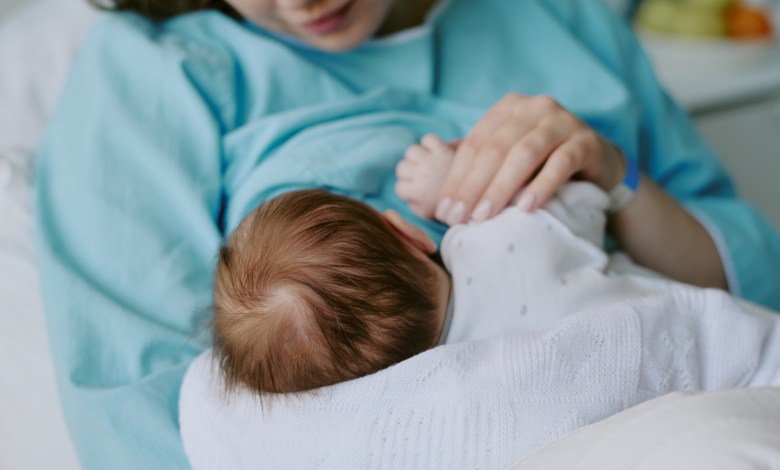 Mother breastfeeding newborn baby in hospital room while holding child gently in arms. Background features hospital equipment and soft focus items creating serene atmosphere
