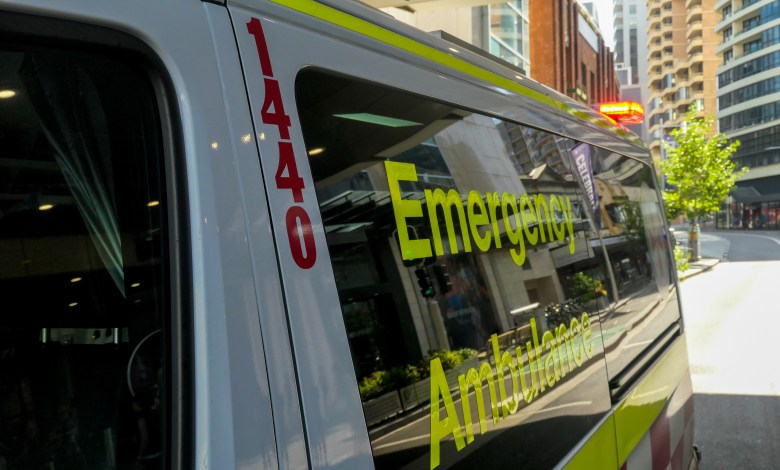 An ambulance of the New South Wales Ambulance Service parked on Oxford Street, Bondi Junction, Sydney. The emergency red light is flashing on the top of the rear door. In the background are commercial and residential buildings. This image was taken on a sunny morning on 11 December 2023.