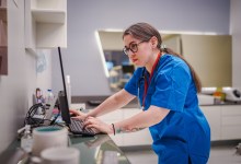 A female healthcare professional efficiently manages patient appointments, using both a computer and a mobile phone at a clinic counter. The scene highlights multitasking, technology use, and administrative duties in a medical environment.