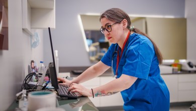 A female healthcare professional efficiently manages patient appointments, using both a computer and a mobile phone at a clinic counter. The scene highlights multitasking, technology use, and administrative duties in a medical environment.