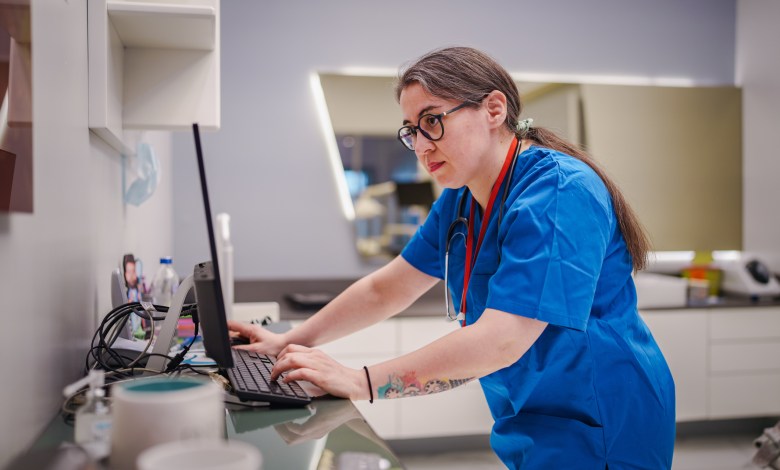 A female healthcare professional efficiently manages patient appointments, using both a computer and a mobile phone at a clinic counter. The scene highlights multitasking, technology use, and administrative duties in a medical environment.