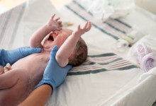 A newborn baby is gently supported by medical staff wearing blue gloves after birth, lying on a hospital bed. The moment captures birth, care, and the start of a family’s new journey.