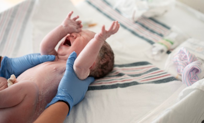 A newborn baby is gently supported by medical staff wearing blue gloves after birth, lying on a hospital bed. The moment captures birth, care, and the start of a family’s new journey.