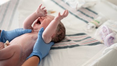 A newborn baby is gently supported by medical staff wearing blue gloves after birth, lying on a hospital bed. The moment captures birth, care, and the start of a family’s new journey.